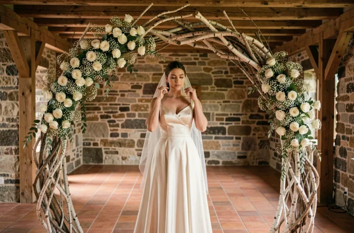 Arche de mariage en branches de bouleau et roses blanches dans une grange en pierre avec mariée en robe de satin ivoire